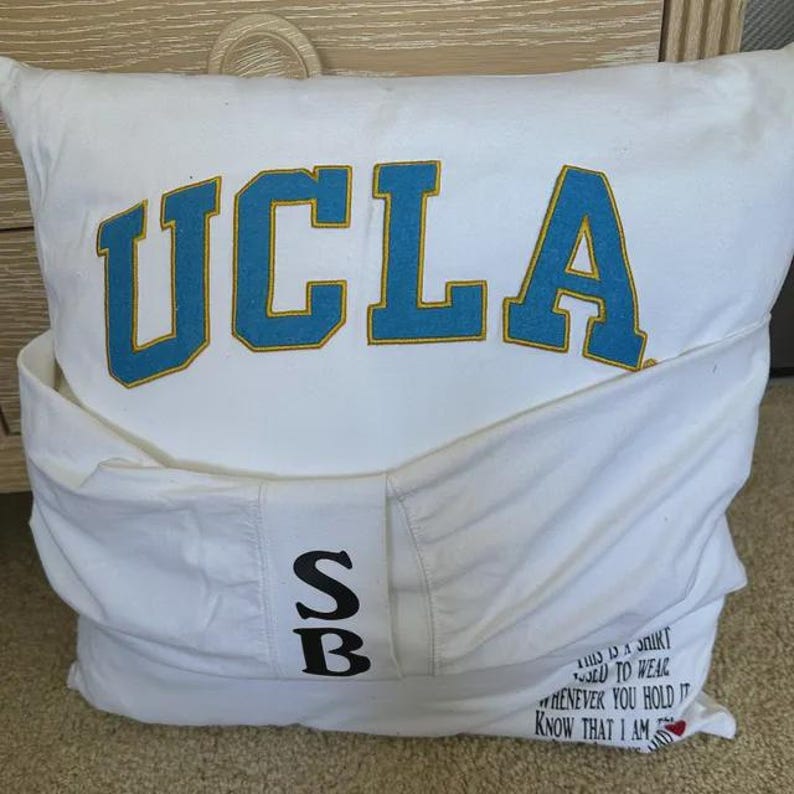 Pode incluir: Travesseiro branco com o texto "UCLA" em letras azuis e amarelas. O travesseiro tem um bolso na frente com as letras "SB" em preto. A parte de tr&aacute;s do travesseiro tem o texto "This is a shirt I used to wear whenever you hold it know that I am with you" em texto preto.