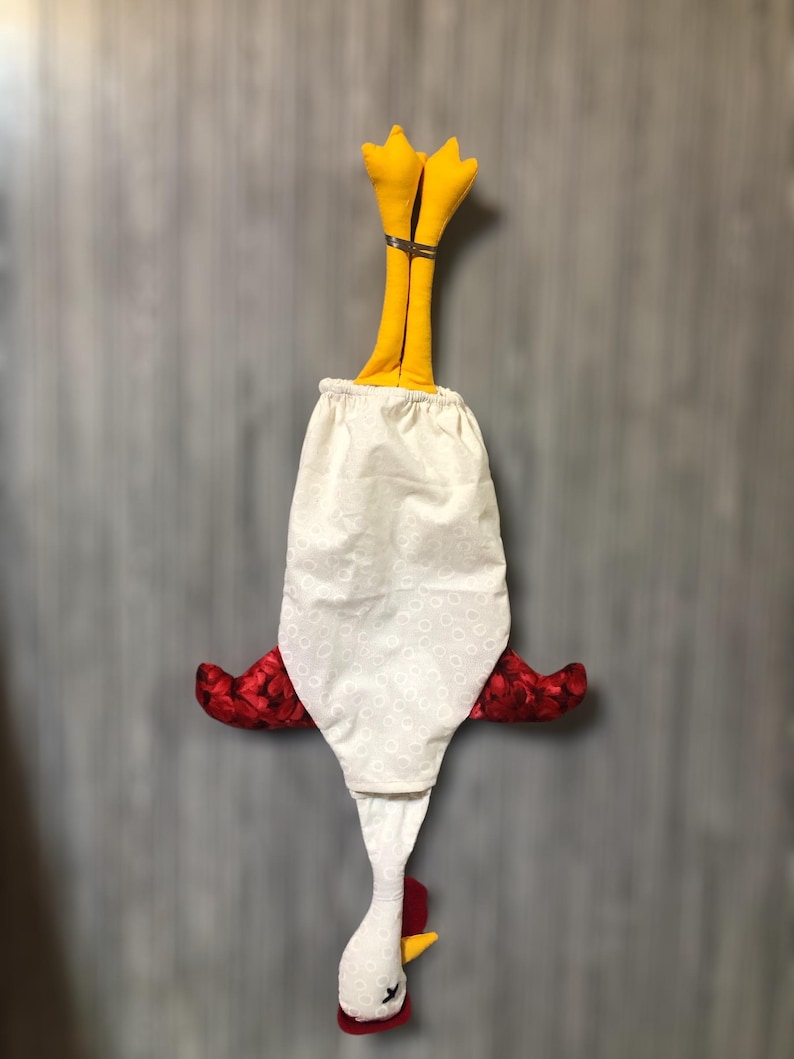 Puede incluir: Un pa&ntilde;o de cocina de tela blanca con forma de pollo, con alas rojas, un pico amarillo y patas amarillas. El pa&ntilde;o est&aacute; colgado boca abajo.