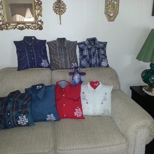 May include: A collection of seven square throw pillows crafted from repurposed button-down shirts in various colors and patterns, including plaid, solid red, and white. Each pillow features a collar, buttons, and a printed memorial message.