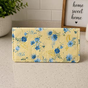 May include: A rectangular fabric checkbook cover featuring a light yellow background with a repeating pattern of blue flowers, green leaves, and small yellow sprigs. The cover has stitched edges and a flat, folded construction.