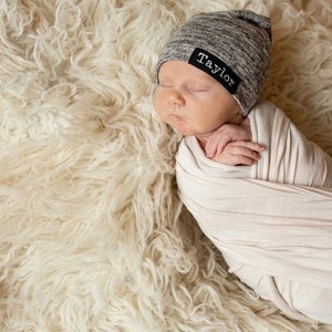 May include: A newborn baby wearing a gray knit beanie with a black label that says "Taylor" is swaddled in a white blanket and lying on a white fluffy rug.