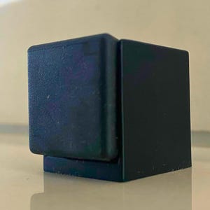 Könnte beinhalten: Ein schwarzer, würfelförmiger Türstopper mit strukturierter Oberfläche auf einer Seite. Der Türstopper ist auf einer kleinen, rechteckigen Basis montiert. Das Objekt besteht aus einem soliden, matten Material und verhindert das Schließen von Türen.