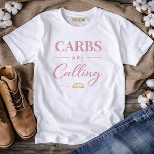 Puede incluir: Camiseta blanca con el texto "CARBS are Calling" en escritura rosa. La camiseta presenta un pequeño gráfico de un cruasán. La camiseta se muestra con botas marrones y vaqueros azules.