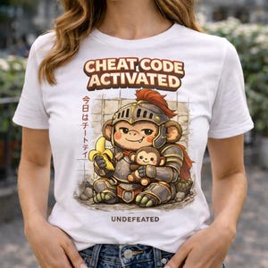 Puede incluir: Camiseta blanca con un mono de dibujos animados con armadura de caballero sosteniendo un plátano y un mono de peluche. El texto incluye "CHEAT CODE ACTIVATED", caracteres japoneses y "UNDEFEATED".