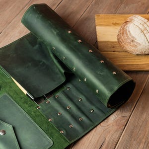以下が含まれることがあります： 金属リベットが付いた、濃い緑色の革製ナイフロールが木製の表面に置かれています。スライスされたパンとナイフが置かれた木製のまな板が隣にあります。ロールには複数のポケットがあります。