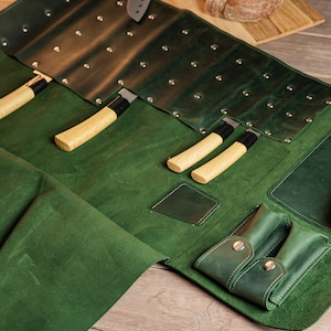 以下が含まれることがあります： 木製の柄と黒い刃が付いたナイフがいくつか入った、緑色の革製ナイフロール。ロールには複数の収納コンパートメントがあります。革は濃い緑色です。ナイフはロールの中にきちんと配置されています。