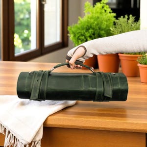 Könnte beinhalten: Eine dunkelgrüne Leder-Rolltasche mit einem stabilen Tragegriff, zwei verstellbaren Schnallenriemen für einen sicheren Verschluss und Metallringen an den Seiten. Die zylindrische Tasche weist sichtbare Nähte und eine matte Oberfläche auf.