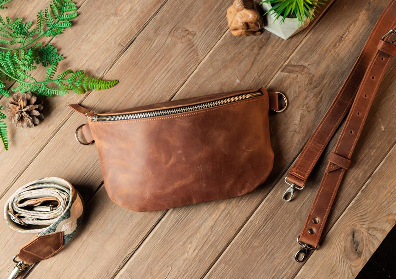 May include: A brown leather fanny pack with a zipper closure and a silver zipper pull. The fanny pack is on a wooden surface with a green plant in the background.