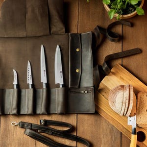 Könnte beinhalten: Eine dunkelbraune Messertasche aus Leder mit vier Fächern für Edelstahl-Küchenmesser, einer Reißverschlusstasche und Messingdruckknöpfen. Ein passender Schulterriemen aus Leder sowie ein Holzschneidebrett mit geschnittenem Brot liegen bei.