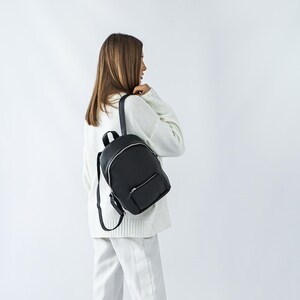 Könnte beinhalten: Ein schwarzer Lederrucksack mit einer kleinen Vordertasche und silbernen Reißverschlüssen. Der Rucksack hat verstellbare Träger und einen oberen Griff. Die Tasche wird über der Schulter getragen. Der Hintergrund ist schlicht weiß.