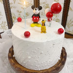 Puede incluir: Una tarta glaseada blanca decorada con figuras de Mickey Mouse y Pluto, adornos de bolas rojas y una vela encendida. La tarta está sobre un soporte de madera y adornada con chispas perladas y globos rojos.