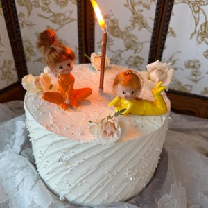 Peut inclure: Un gâteau rond blanc avec un glaçage texturé et des perles blanches. Deux petites poupées aux cheveux roux, l'une en orange et l'autre en jaune, sont posées dessus. Une bougie allumée se trouve entre elles, entourée de trois roses blanches.