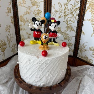 Può includere: Una torta bianca glassata decorata con statuine di Topolino, Minnie e Pluto. La torta è adornata con decorazioni sferiche rosse e piccole perle bianche. La torta poggia su un alzatina di legno.