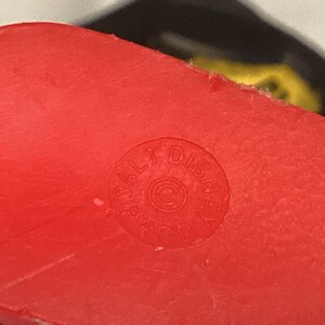 May include: Close-up of a bright red plastic object with a circular stamp that reads "WALT DISNEY PROD" around a copyright symbol. The object has a slightly textured surface.