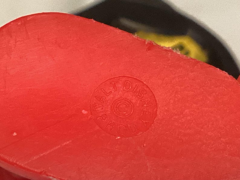 May include: Close-up of a bright red plastic object with a circular stamp that reads "WALT DISNEY PROD" around a copyright symbol. The object has a slightly textured surface.