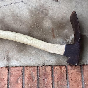May include: A garden hoe with a curved, dark metal blade and a light-colored wooden handle. The tool rests on a concrete surface with a brick border, showcasing its design and construction for gardening tasks.