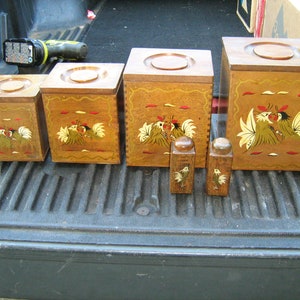 May include: Set of four wooden canisters with rooster designs painted on them. The canisters are different sizes and have lids. Two smaller wooden containers are also included.