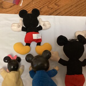 Puede incluir: Una colecci&oacute;n de peluches de Mickey Mouse. Los juguetes tienen cabezas negras con orejas grandes, guantes blancos, pantalones cortos rojos y zapatos amarillos. Algunos juguetes tienen una camisa roja y pantalones azules. Los juguetes est&aacute;n sobre una superficie blanca.
