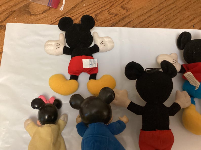 Puede incluir: Una colecci&oacute;n de peluches de Mickey Mouse. Los juguetes tienen cabezas negras con orejas grandes, guantes blancos, pantalones cortos rojos y zapatos amarillos. Algunos juguetes tienen una camisa roja y pantalones azules. Los juguetes est&aacute;n sobre una superficie blanca.