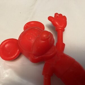 May include: A red plastic figurine of a cartoon mouse with large ears, a round head, and a raised hand. The figure is in a playful pose, with its arm extended upwards. The toy is made of solid red plastic.