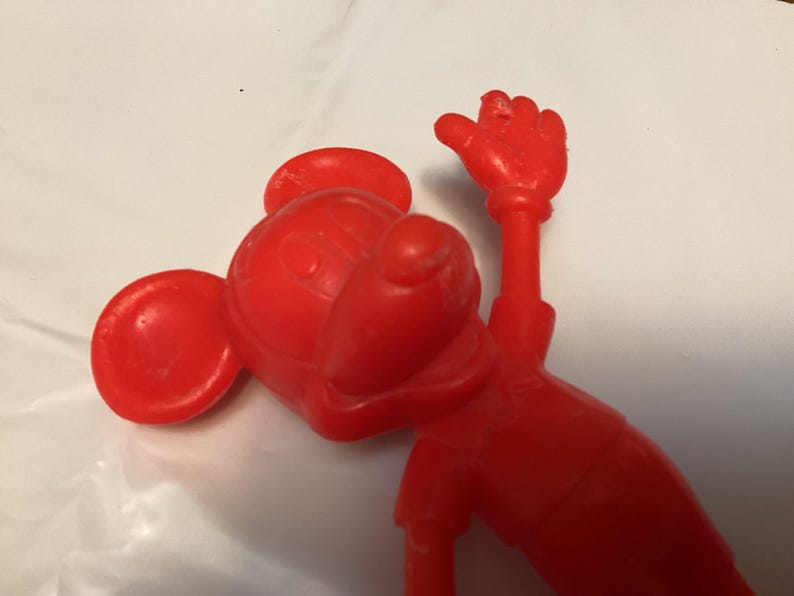 May include: A red plastic figurine of a cartoon mouse with large ears, a round head, and a raised hand. The figure is in a playful pose, with its arm extended upwards. The toy is made of solid red plastic.