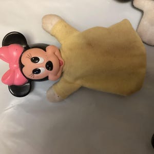 Puede incluir: Una marioneta de mano de Minnie Mouse con cabeza negra, lazo rosa y cuerpo amarillo. La cara de la marioneta es de pl&aacute;stico, con ojos negros y boca roja. El cuerpo es de tela amarilla descolorida.