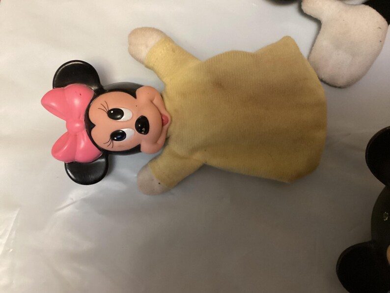 Puede incluir: Una marioneta de mano de Minnie Mouse con cabeza negra, lazo rosa y cuerpo amarillo. La cara de la marioneta es de pl&aacute;stico, con ojos negros y boca roja. El cuerpo es de tela amarilla descolorida.