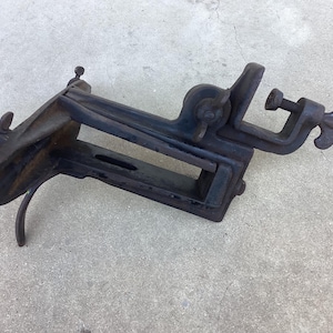 May include: An antique, cast iron tool with a dark, weathered finish. The tool features a clamp, a screw, and a lever, suggesting it was used for holding or shaping materials. The design is robust and industrial, indicative of its age and purpose.