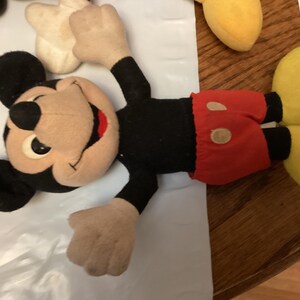 Puede incluir: Un peluche vintage de Mickey Mouse. El peluche presenta un cuerpo, orejas y brazos negros, con una cara, manos y pies de color beige. Mickey lleva pantalones cortos rojos con dos botones blancos y zapatos amarillos. El juguete est&aacute; sobre una superficie blanca.