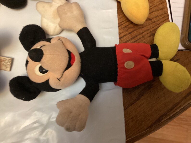 Puede incluir: Un peluche vintage de Mickey Mouse. El peluche presenta un cuerpo, orejas y brazos negros, con una cara, manos y pies de color beige. Mickey lleva pantalones cortos rojos con dos botones blancos y zapatos amarillos. El juguete est&aacute; sobre una superficie blanca.