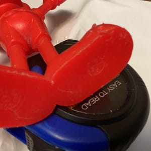 May include: A red plastic toy figure with large feet is positioned over a black and blue measuring tape. The text "EASY TO READ" is visible on the tape measure. The toy figure is in a playful pose.