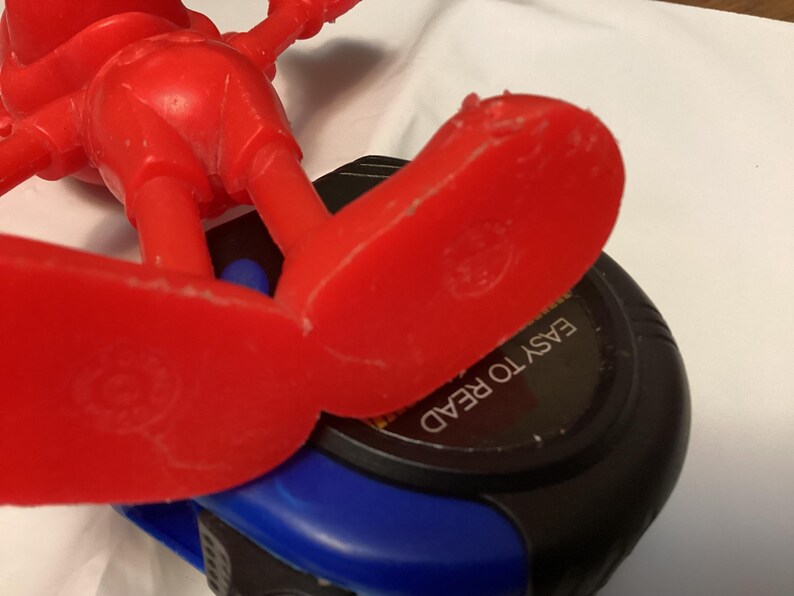 May include: A red plastic toy figure with large feet is positioned over a black and blue measuring tape. The text "EASY TO READ" is visible on the tape measure. The toy figure is in a playful pose.