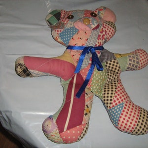 May include: A patchwork teddy bear with a blue ribbon tied around its neck. The bear is made of various colorful fabrics, including red, pink, green, blue, and yellow.