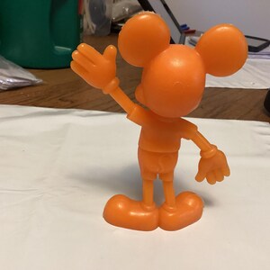 May include: Orange plastic figurine of a mouse-like character with large round ears, a smiling face, and gloved hands. The figure is posed with one arm raised in a wave. The character is wearing shorts and shoes.