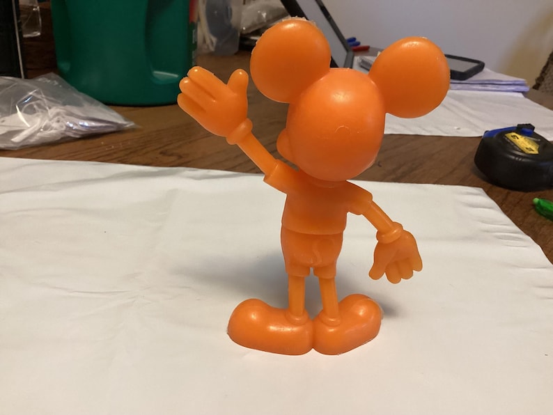 May include: Orange plastic figurine of a mouse-like character with large round ears, a smiling face, and gloved hands. The figure is posed with one arm raised in a wave. The character is wearing shorts and shoes.