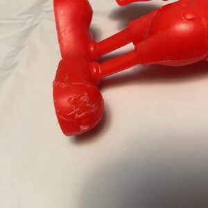 May include: A close-up of a red plastic toy figure's leg and foot. The foot has visible scratches and imperfections. The figure appears to be part of a larger toy, possibly a character from a cartoon or children's show.