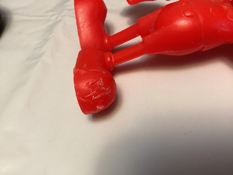 May include: A close-up of a red plastic toy figure's leg and foot. The foot has visible scratches and imperfections. The figure appears to be part of a larger toy, possibly a character from a cartoon or children's show.
