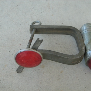 May include: A vintage metal bicycle tail light with a red lens and a silver metal clamp. The light has a round red lens and a silver metal body.