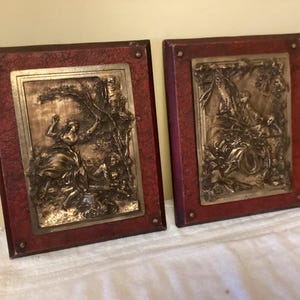 May include: Two framed wall decor pieces with raised, silver-toned, embossed images of people in a garden setting. The frames are dark red with a textured surface.