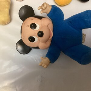 Puede incluir: Un peluche vintage de Mickey Mouse con un mono azul. El juguete tiene una cara pintada con grandes ojos negros y una nariz negra. Las orejas son negras y las manos y los pies son de color marr&oacute;n claro.