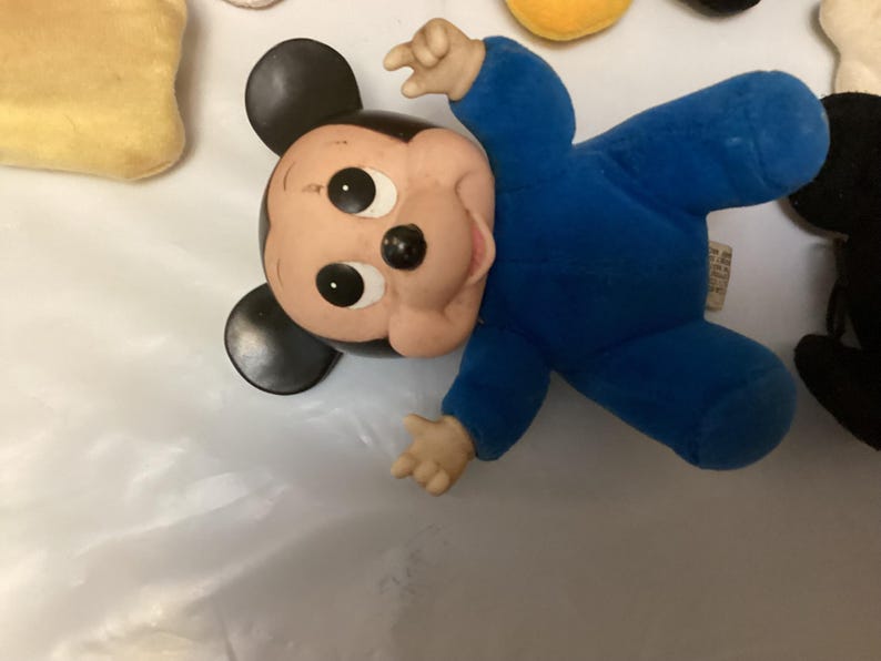 Puede incluir: Un peluche vintage de Mickey Mouse con un mono azul. El juguete tiene una cara pintada con grandes ojos negros y una nariz negra. Las orejas son negras y las manos y los pies son de color marr&oacute;n claro.