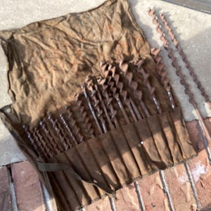 May include: A set of vintage drill bits in a brown canvas tool roll. The bits are various sizes and made of metal. The tool roll is open, displaying the bits, and is lying on a brick and concrete surface.