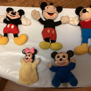 Puede incluir: Cinco mu&ntilde;ecos de peluche de Mickey Mouse y Minnie Mouse. Los mu&ntilde;ecos tienen orejas negras, guantes blancos y zapatos amarillos. Mickey lleva pantalones cortos rojos y una camisa roja. Minnie lleva un lazo rosa y un vestido amarillo. Los mu&ntilde;ecos est&aacute;n sobre una superficie blanca.