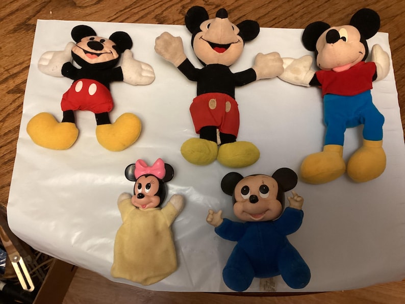 Puede incluir: Cinco mu&ntilde;ecos de peluche de Mickey Mouse y Minnie Mouse. Los mu&ntilde;ecos tienen orejas negras, guantes blancos y zapatos amarillos. Mickey lleva pantalones cortos rojos y una camisa roja. Minnie lleva un lazo rosa y un vestido amarillo. Los mu&ntilde;ecos est&aacute;n sobre una superficie blanca.