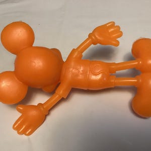 May include: Orange plastic toy figure of a cartoon character, lying on its back. The figure has a round head with two smaller ear-like circles, arms, legs, and large orange shoes. The figure is a solid orange color.