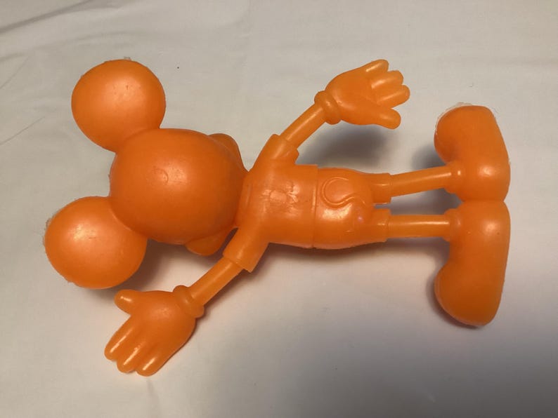 May include: Orange plastic toy figure of a cartoon character, lying on its back. The figure has a round head with two smaller ear-like circles, arms, legs, and large orange shoes. The figure is a solid orange color.
