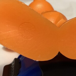 May include: Close-up of an orange plastic toy's feet, with a circular marking on the sole. The toy has a blue and black base, and another orange piece is visible in the background. The toy appears to be vintage.