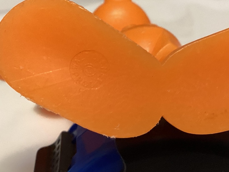 May include: Close-up of an orange plastic toy's feet, with a circular marking on the sole. The toy has a blue and black base, and another orange piece is visible in the background. The toy appears to be vintage.