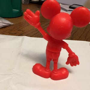 May include: A solid red figurine of a cartoon mouse with large round ears, a round head, and a smiling face. The mouse is posed with one arm raised in a waving gesture. The figurine is standing on a white surface.