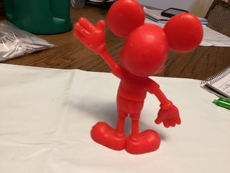 May include: A solid red figurine of a cartoon mouse with large round ears, a round head, and a smiling face. The mouse is posed with one arm raised in a waving gesture. The figurine is standing on a white surface.